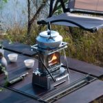 Picknick utomhus med gasbrännare och aluminiumkanna på ett träbord, omgiven av natur, perfekt för campingäventyr eller utomhusutflykter.