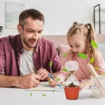 En far och hans dotter genomför ett vetenskapligt experiment hemma med ett terrarium, ett provrör och gröna blad, vilket främjar lärande genom lek.