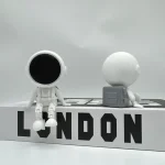 Figurer av vetenskapliga utforskare i minimalistisk stil med hjälm och glob, på en plattform med ordet "LONDON" i svarta bokstäver.
