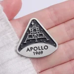 Märke i form av en triangel som föreställer månmodulen Apollo 1969, med detaljer i relief och inskriptionen "APOLLO 1969", perfekt för samlare och rymdentusiaster.