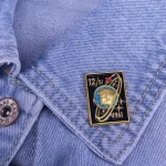 Kravattklämma i metall med rymdmotiv och datum 1961, fäst på en ljusblå denimskjorta, vilket ger en vintage och vetenskaplig stil till outfiten.