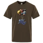 Brun t-shirt med mönster av en astronaut som svävar och bär en ballong formad av flera färgglada planeter, vilket skapar en lekfull och fantasifull rymdscen.