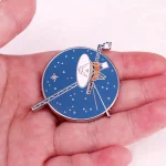 En brosch som föreställer en rymdstation med antenner och solpaneler, idealisk för astronomi- och vetenskapsdekorationentusiaster.