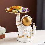 Astronautfigur i vit keramik med gyllene hjälm, som håller en bricka fylld med färgglada godisar, originaldekoration för ett tematiskt utrymme eller en samling av vetenskapliga objekt.