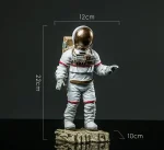 Miniatyrfigur av astronaut med gyllene hjälm, 22 cm hög, 12 cm bred och 10 cm djup, placerad på en stenig bas, idealisk för vetenskaplig dekoration.