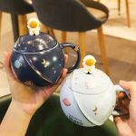 Kosmisk kopp med lock och astronautfigur, dekorerad med mönster av planeter, stjärnor och månen, idealisk för astronomientusiaster och vetenskaplig dekoration.