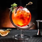 Färgglad cocktail i ett originellt glas med mynta och citronskiva, presenterad på en svart matta med baraccessoarer i bakgrunden.