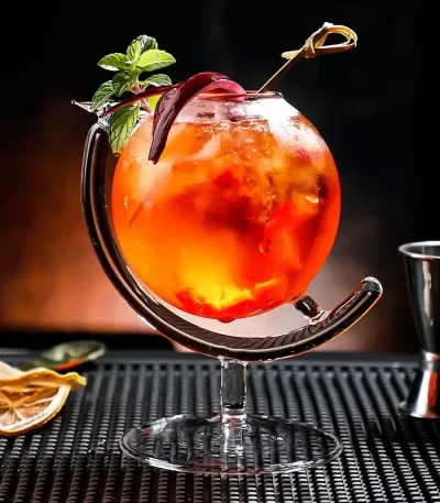 Färgglad cocktail i ett originellt glas med mynta och citronskiva, presenterad på en svart matta med baraccessoarer i bakgrunden.