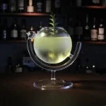 Lysande cocktail i en vetenskaplig burk med en flytande grön växt, presenterad på ett glasstöd, barstämning med hyllor av flaskor i bakgrunden.