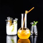 Laboratorievase med en orange dryck och ett sugrör, tillsammans med andra glaslaboratorietillbehör, på svart bakgrund.