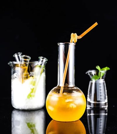 Laboratorievase med en orange dryck och ett sugrör, tillsammans med andra glaslaboratorietillbehör, på svart bakgrund.