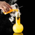 Genomförande av ett vetenskapligt experiment med rykande orange vätska i ett laboratorium, med en mätkolv och ett provrör, mot en svart bakgrund.