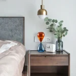 Orange och blå glasvas på ett modernt nattduksbord med grön växt och hängande gyllene lampa.
