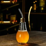 Dekorativ glödlampa i form av en flaska med orange dryck, utrustad med ett metallrör, på en träyta i en mörk och modern miljö.