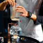 Glaspipett som hanteras av en hand under ett vetenskapligt experiment, med en laboratorieballong som innehåller en transparent vätska, vilket illustrerar precision och teknik inom kemi.
