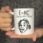 Mugg med den berömda ekvationen av Einstein E=MC² och en humoristisk illustration av Einstein med tungan ute, idealisk för vetenskapsentusiaster och geekinredning.