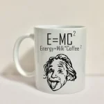 Vit mugg med formeln E=MC², kopplad till "Energy=Milk*Coffee²" och en humoristisk teckning av Einstein med tungan ute, som illustrerar en rolig blandning av vetenskap och kaffe.
