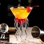 Vetenskapligt cocktailglas med dekoration i form av tentakler i transparent glas, fyllt med en gul-orange cocktail med körsbär, modern baratmosfär.