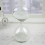 Timglas i glas med vit sand, symbol för tidens gång, placerat på en ljus yta med en minimalistisk bakgrund.