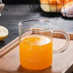 Kemiglas i transparent glas fyllt med en orange vätska, placerad på en träplatta, med frukter i bakgrunden.