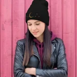 Ung kvinna som bär en svart mössa med mån- och stjärnmotiv, klädd i en svart läderjacka och en lila hoodie, poserar framför en rosa trävägg.