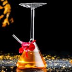 Laboratoriebägare med hängande cocktailglas, innehållande en orange vätska, dekorerad med en röd blomma, på en svart yta med gyllene ljusglimtar.