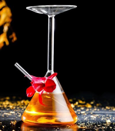Laboratoriebägare med hängande cocktailglas, innehållande en orange vätska, dekorerad med en röd blomma, på en svart yta med gyllene ljusglimtar.