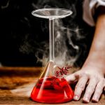 Laboratorieflaska i glas med en röd rykande vätska, placerad på en träyta, med en hand bredvid, som illustrerar ett vetenskapligt experiment.