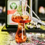 Originellt cocktailglas i form av en laboratorieballong, dekorerat med granbarr, perfekt för festdekoration eller vetenskapligt evenemang.