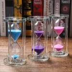 Färgglatt glas-timglas med tre rör som innehåller blå, lila och rosa sand, placerat på en träyta med böcker i bakgrunden.