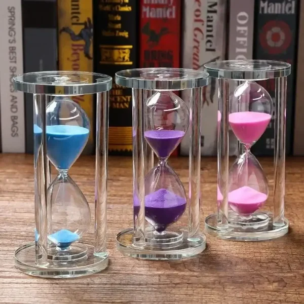 Färgglatt glas-timglas med tre rör som innehåller blå, lila och rosa sand, placerat på en träyta med böcker i bakgrunden.