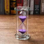Glas-timglas med lila sand, symboliserar tidens gång, placerad på en träyta med en bokhylla i bakgrunden.