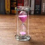 Glas-timglas med ljusrosa sand, symboliserar tidens gång, placerad på en träyta med en bokhylla i bakgrunden.