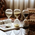 Tre timglas i glas med vit, gul och svart sand, placerade på ett träbord i en vintageinredning, som illustrerar tidens mätning med elegans.