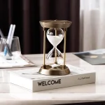 Dekorativ sandtopp i mässing och glas, placerad på en bok med titeln "WELCOME" i en kontors- eller vardagsrumsmiljö.