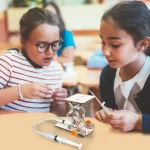 Två unga flickor utforskar en utbildningsrobot under en klassaktivitet, vilket illustrerar lekfullt lärande om teknik och robotik för barn.