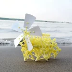 Miniveteckningsfordon i plast i gult med vit propeller, placerat på sanden nära havet, som illustrerar ett original och pedagogiskt vetenskapligt dekorationsobjekt.