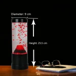 Säpbubbelvas med röd LED-lampa, höjd 25,5 cm och diameter 9 cm, placerad på ett träbord med glasögon och en anteckningsbok bredvid, svart bakgrund.